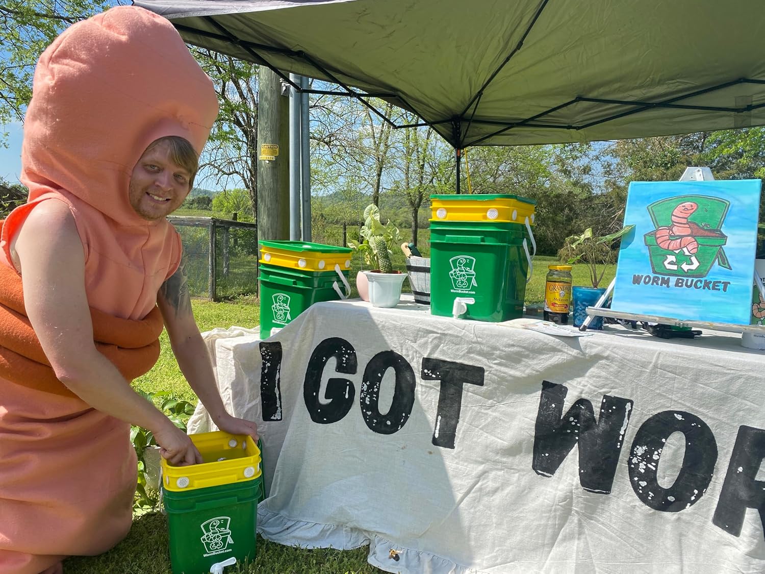 Person in a worm costume interacting with green containers under a tent with 'I GOT WORMS text.